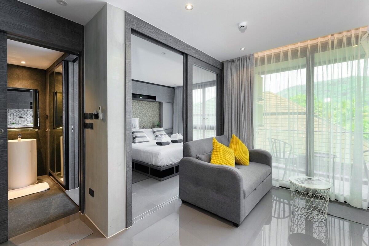 Ein modernes Hotelzimmer mit einem Bett, einem grauen Sofa mit gelben Kissen, einem kleinen runden Tisch und einem großen Fenster mit Vorhängen.