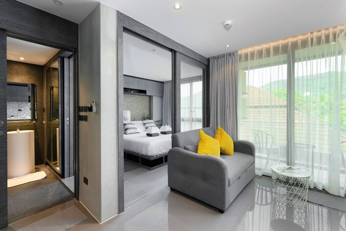 A modern hotel room with a bed, a grey sofa with yellow pillows, a small round table, and a large window with curtains.