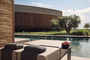 A poolside lounge area with a modern building in the background, a clear view of the pool, and a bowl of fruit on the lounge. - Zelia Halkidiki (Nea Propontida)