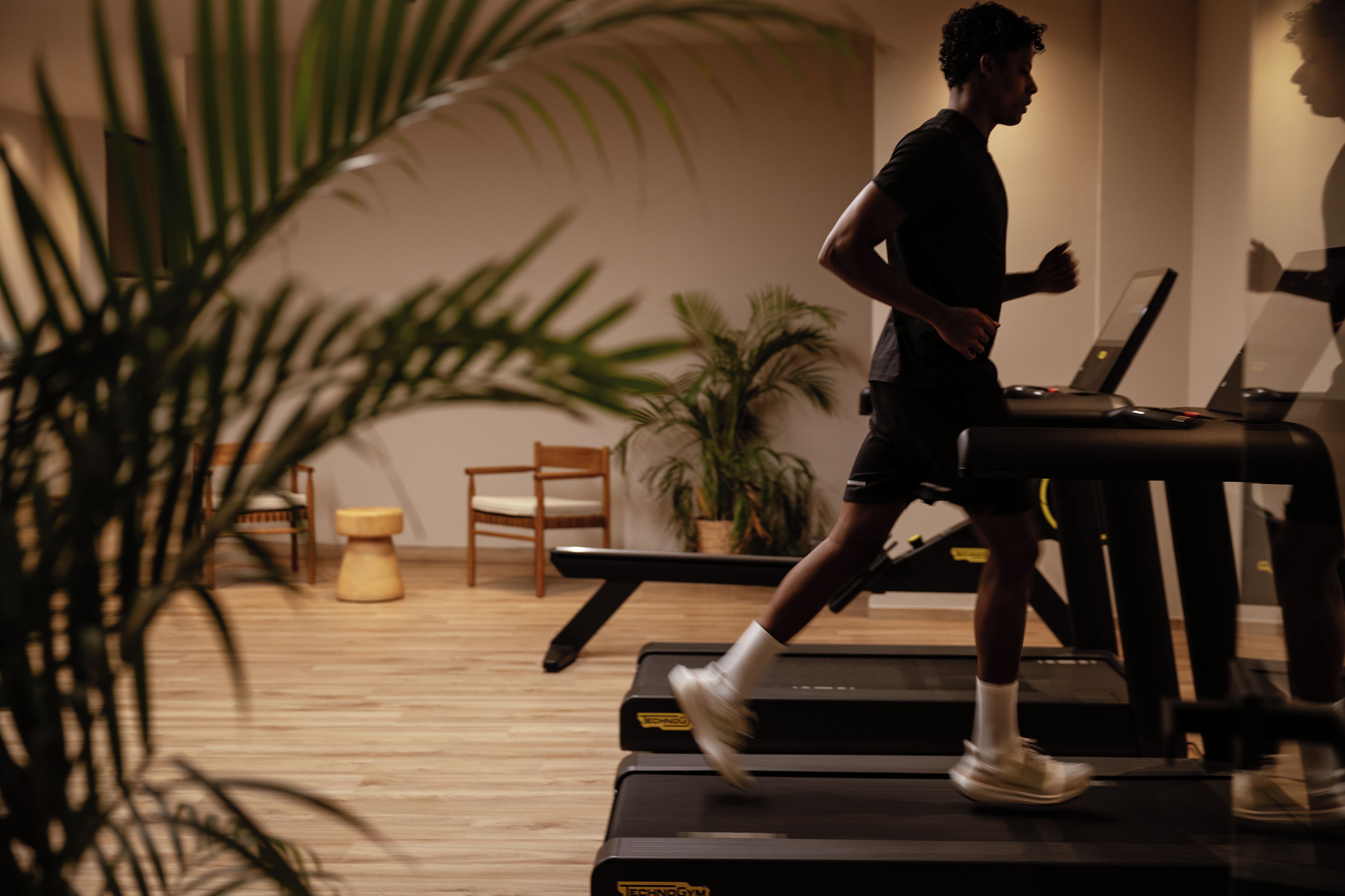 Une personne court sur un tapis de course dans une pièce dont le sol est en bois, et qui contient une chaise, une petite table et une plante en pot.