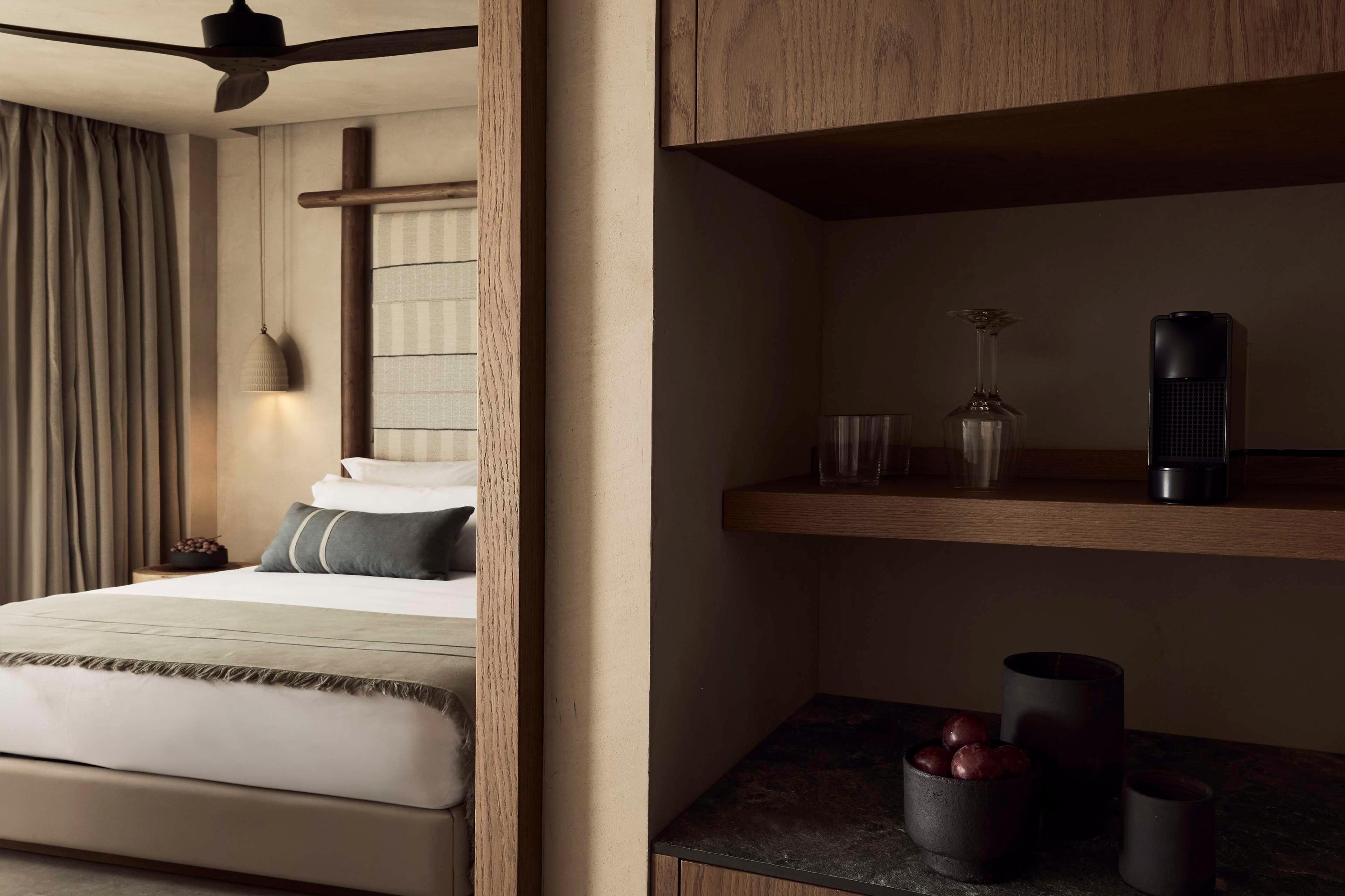 A hotel room with a bed, a bedside table with a vase and a bottle, and a wooden shelf with a wine glass and a speaker.