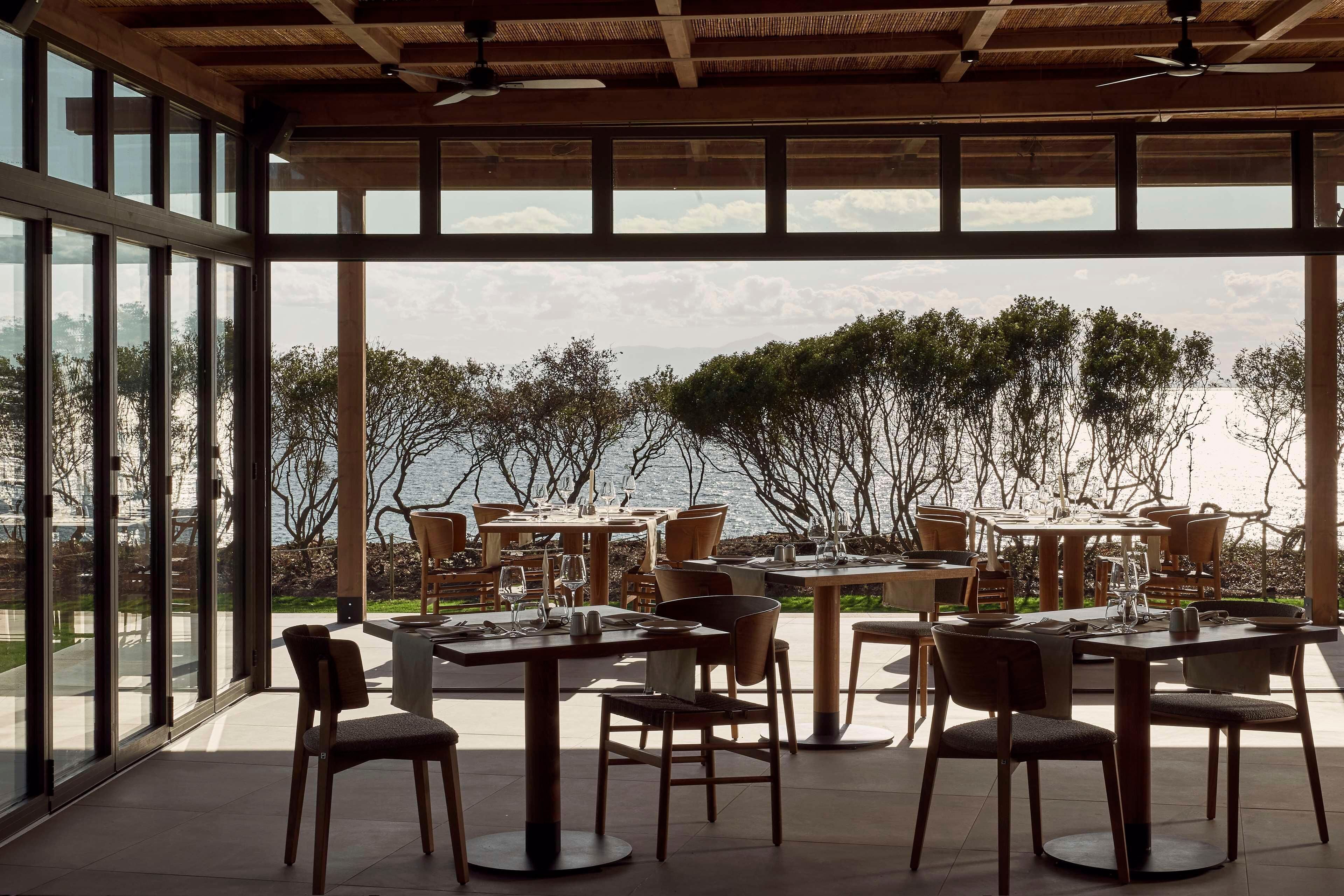 An outdoor dining area with tables and chairs, large windows, and a view of the sea.