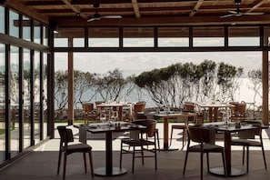 Un espace extérieur aménagé pour les repas, avec des tables et des chaises, de grandes fenêtres et une vue sur la mer.