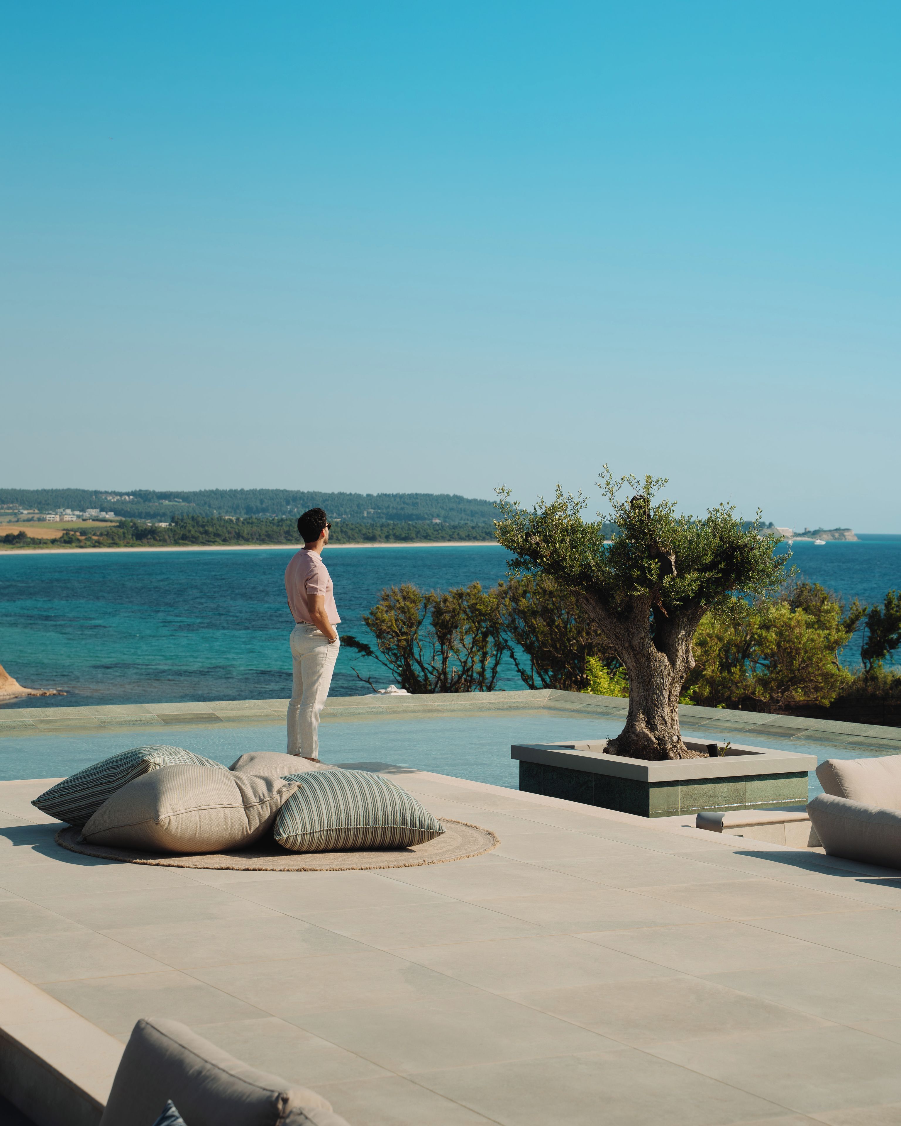 A person standing by a pool overlooking a beach with a tree and cushions.