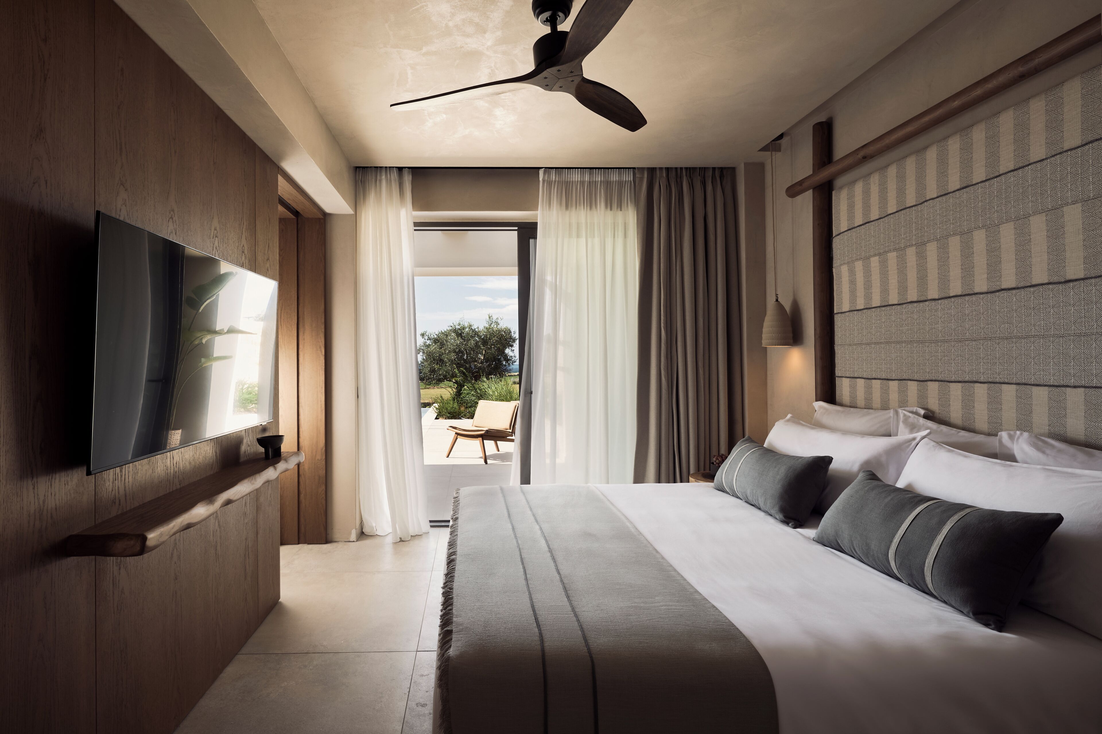 A modern hotel room with a large bed, a flat-screen TV, a ceiling fan, and a view of the outdoors.
