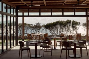 An outdoor dining area with tables and chairs, large windows, and a view of the sea. - Zelia Halkidiki (Nea Propontida)