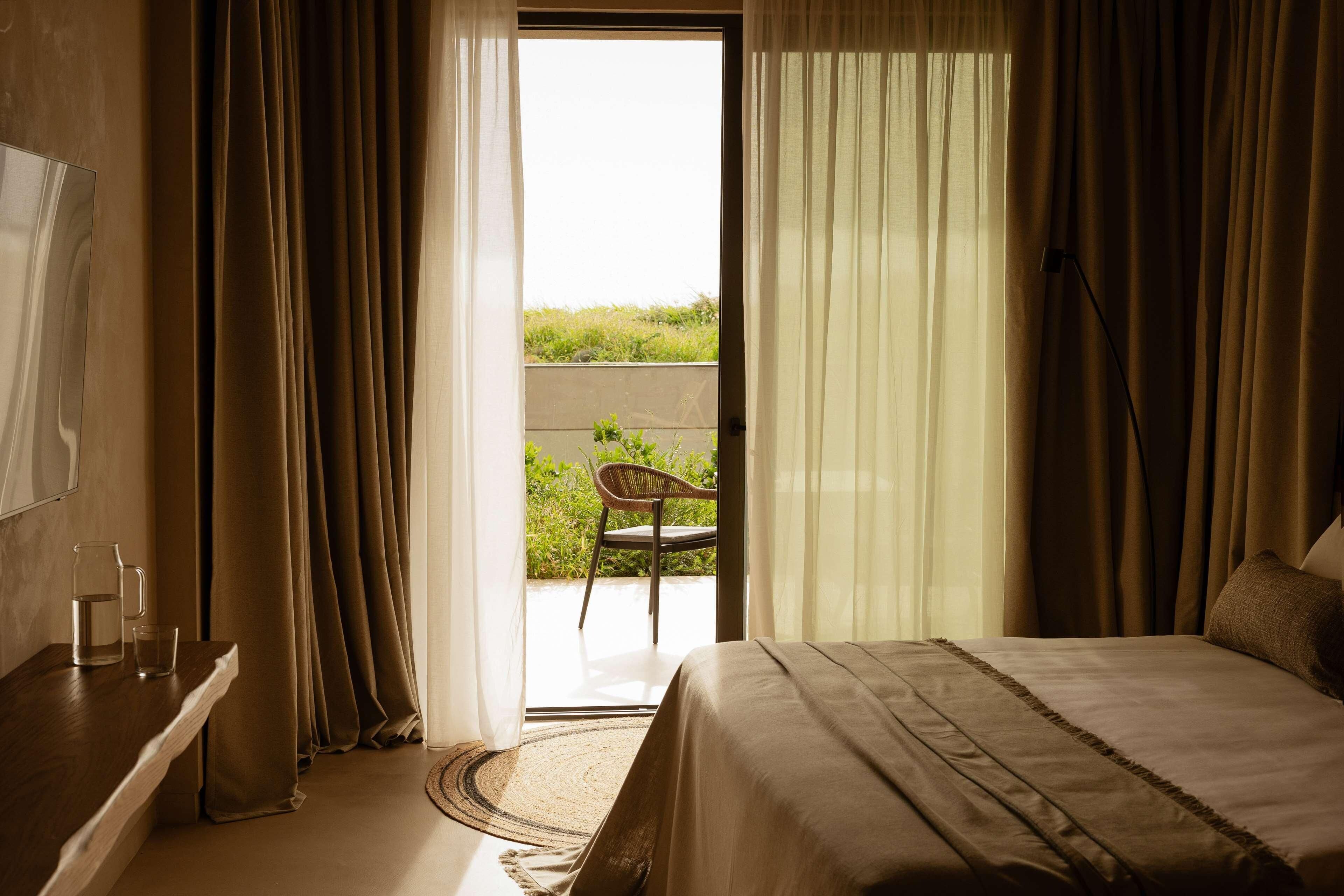 A bedroom with a bed, a chair outside, and a view of greenery.