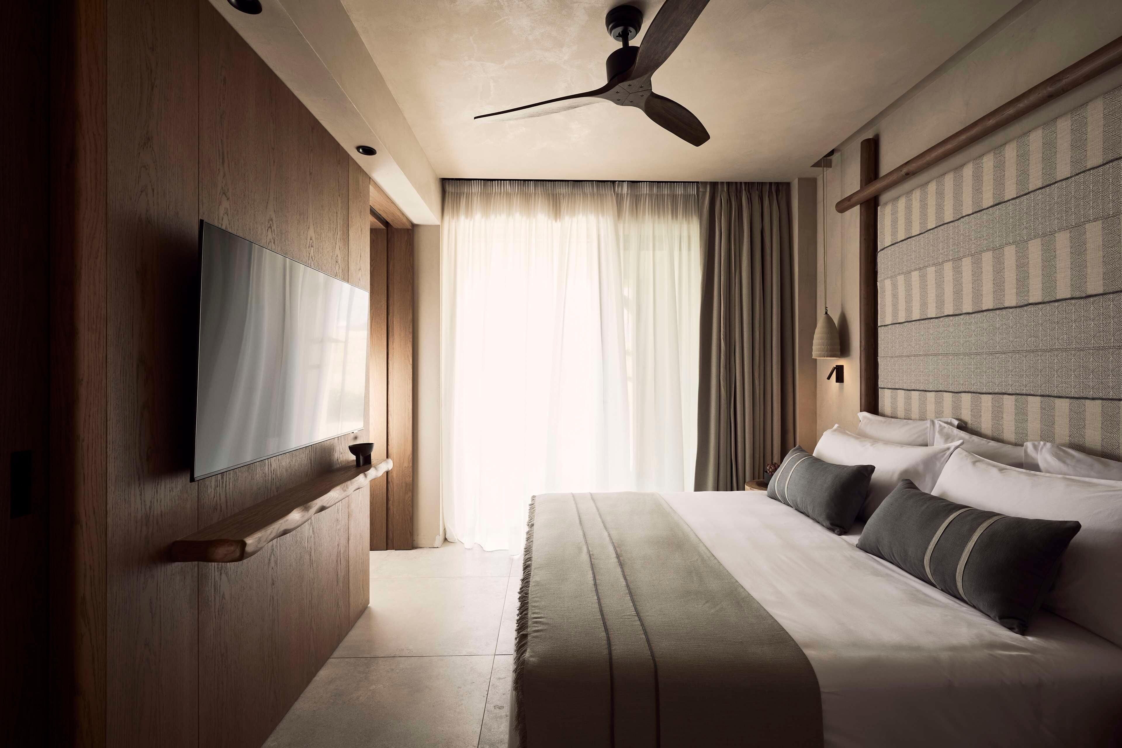 A modern hotel room with a large bed, a ceiling fan, a mounted TV, and a large window with curtains.