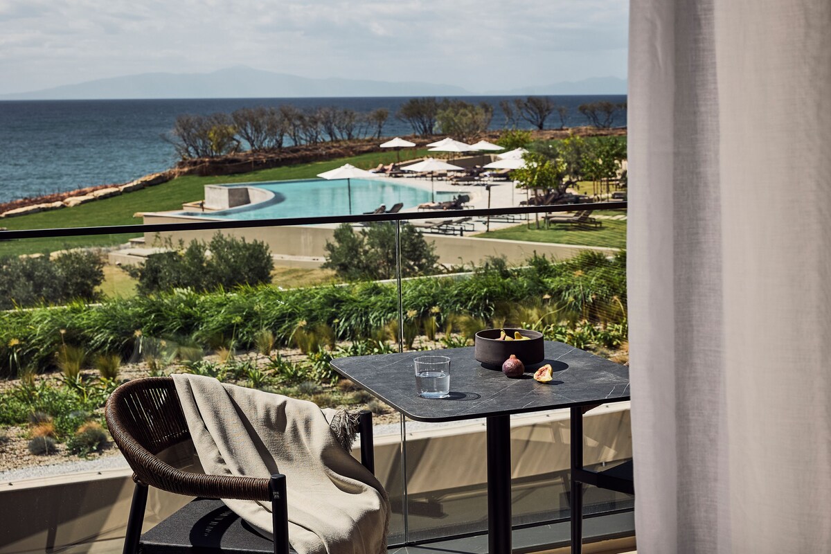 A balcony with a view of a pool, lounge chairs, and a dining table with a glass and fruit.