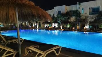 Una piscina nocturna con sillones de descanso y un toldo de paja.
