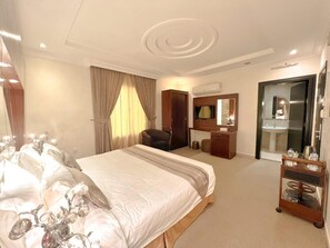 Ein Hotelzimmer mit einem großen Bett, einem Schreibtisch, einem Stuhl, einem Frisiertisch mit Spiegel und einem durch eine offene Tür einsehbaren Badezimmer.