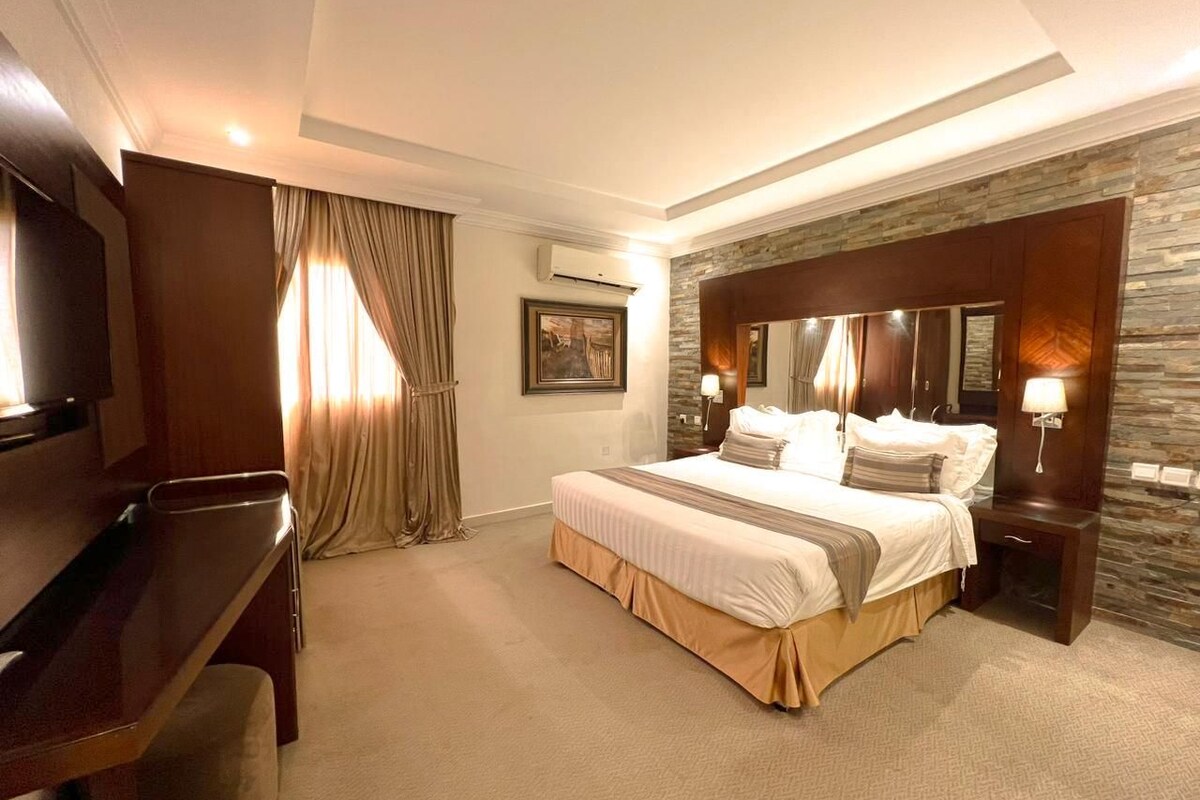 Habitación de hotel con una cama grande, un escritorio, una silla, un equipo de aire acondicionado y una pared decorativa de piedra.