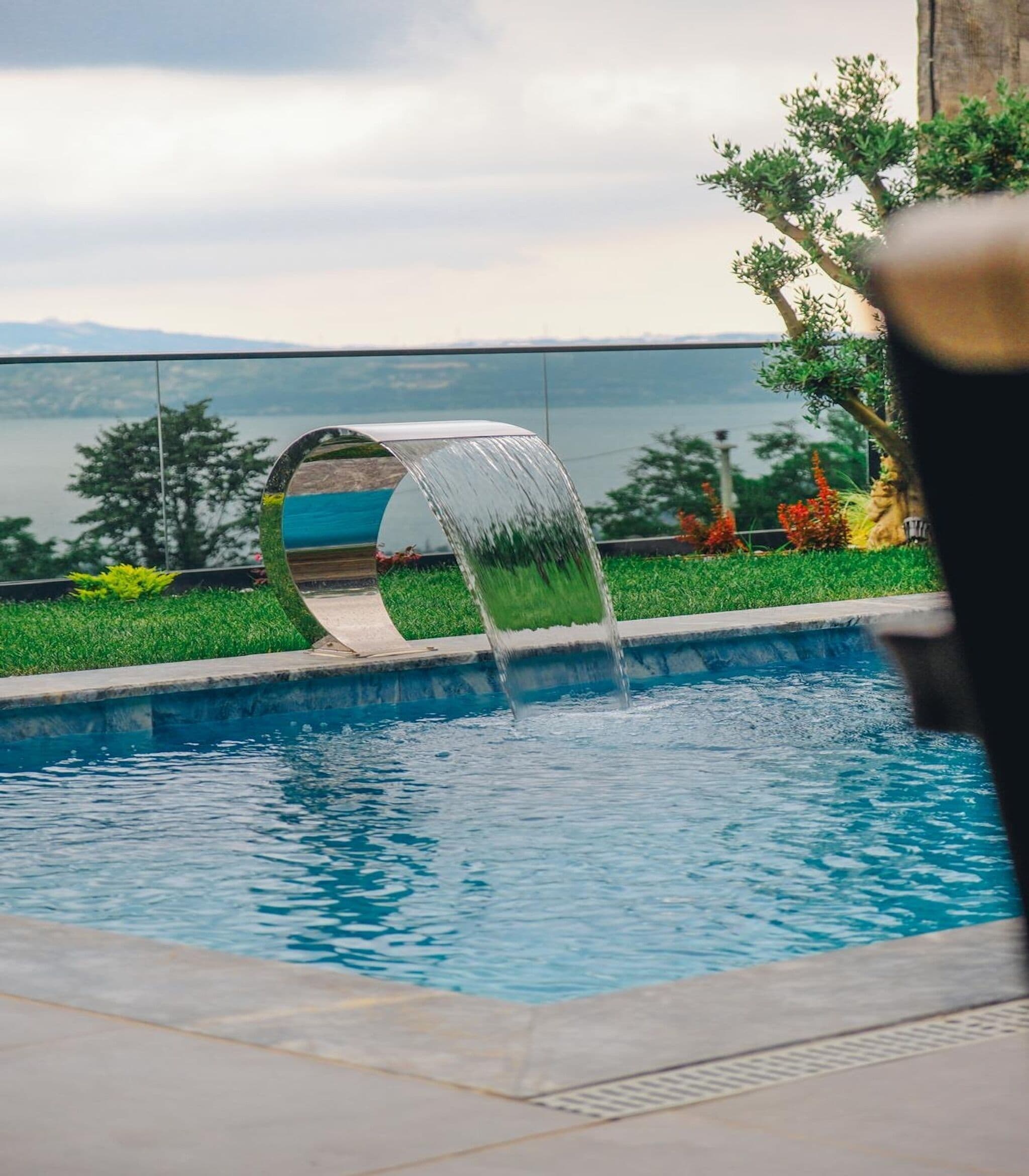 A pool with a water feature, a clear view of the sky, and a landscape with trees and plants.