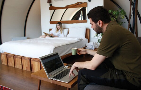 A room with a bed, a laptop on a wooden table, and a person sitting on the floor.