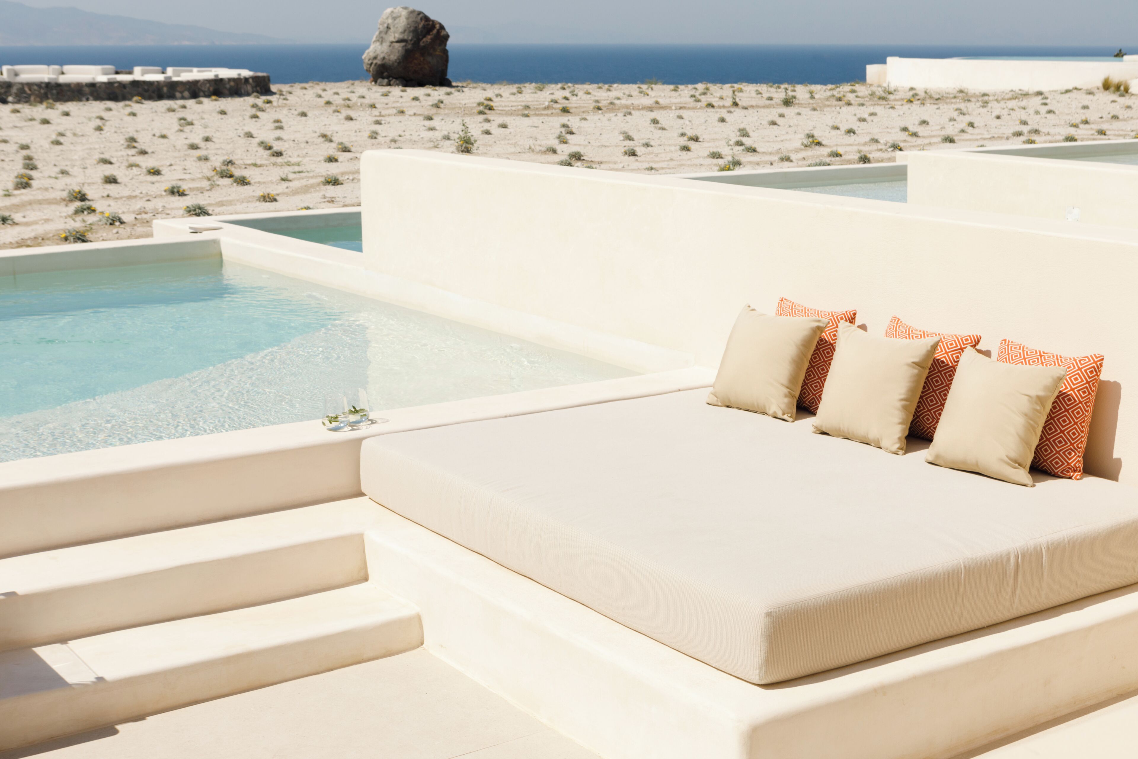 A poolside seating area with steps leading to the water, a view of the beach, and a large rock in the background.