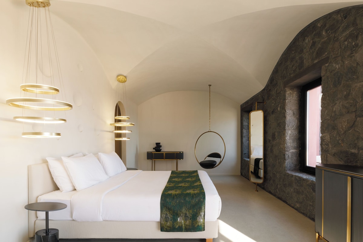 Ein modernes Schlafzimmer mit einem großen Bett, einem Hängekorbsessel, einem runden Spiegel und einer Steinwand.