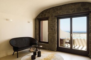Ein Zimmer mit einem schwarzen Sessel, einem kleinen runden Tisch und einer großen Schiebeglasdoor, die zu einem Sitzbereich im Freien mit Blick auf den Strand führt.