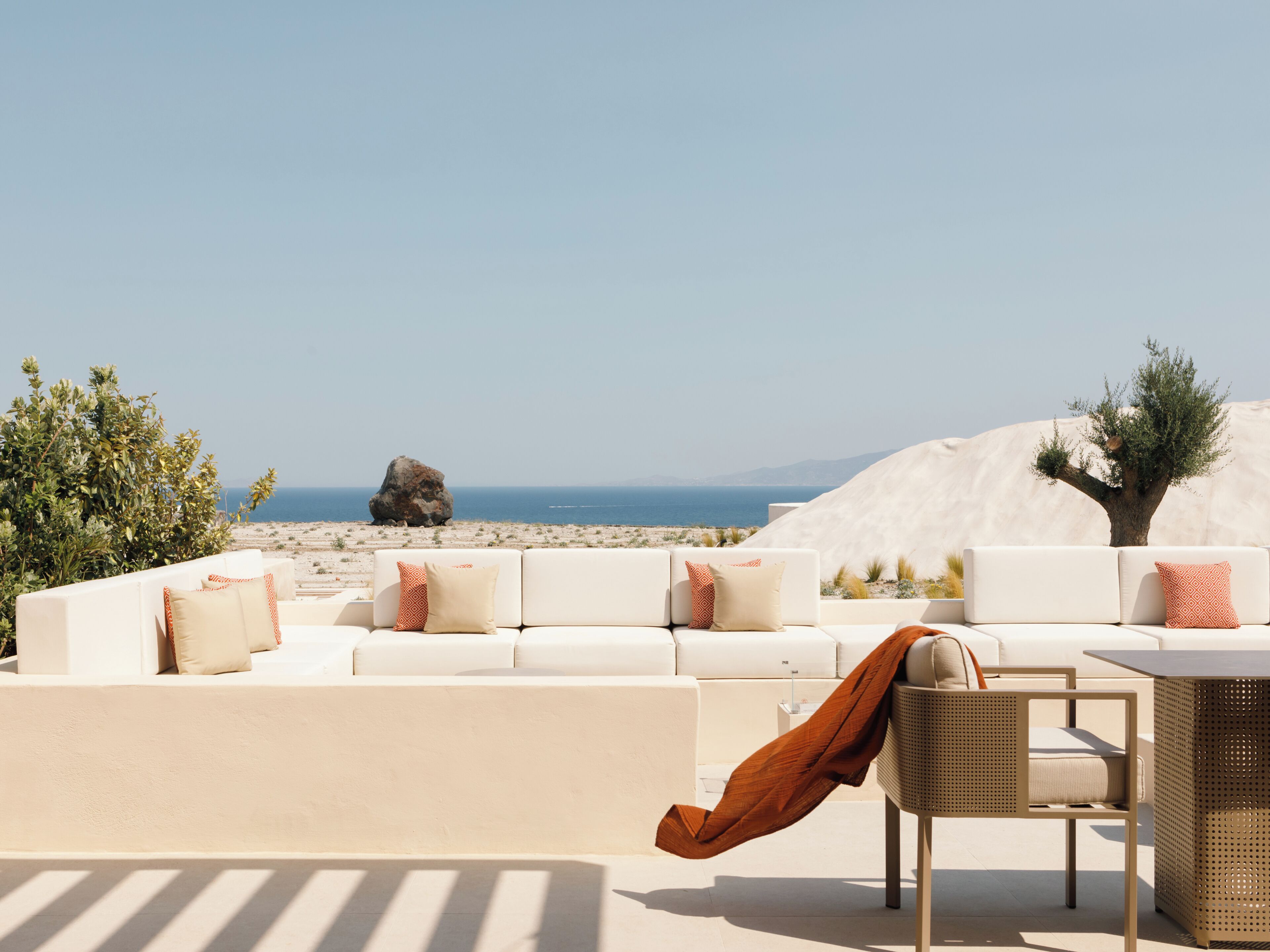 A sunlit outdoor seating area with a white sofa and chairs, a table, and a view of the sea and mountains.
