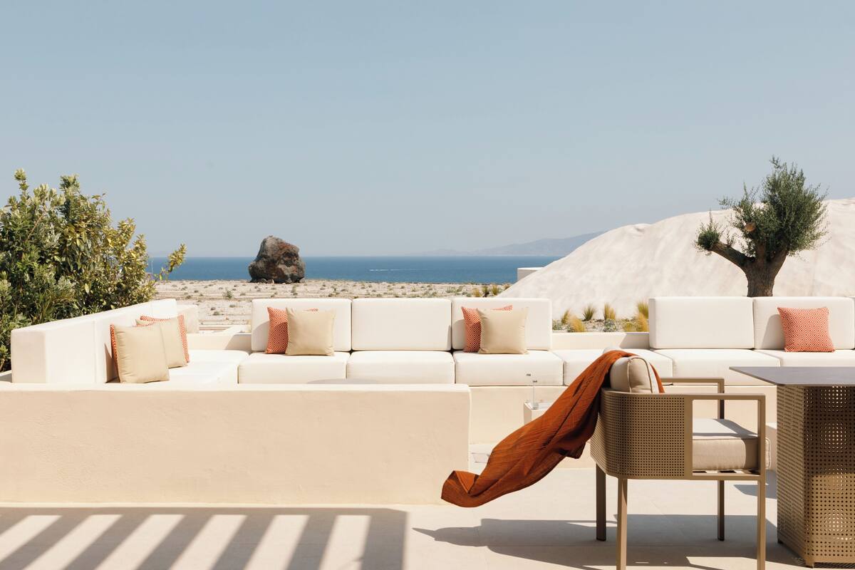 A sunlit outdoor seating area with a white sofa and chairs, a table, and a view of the sea and mountains.