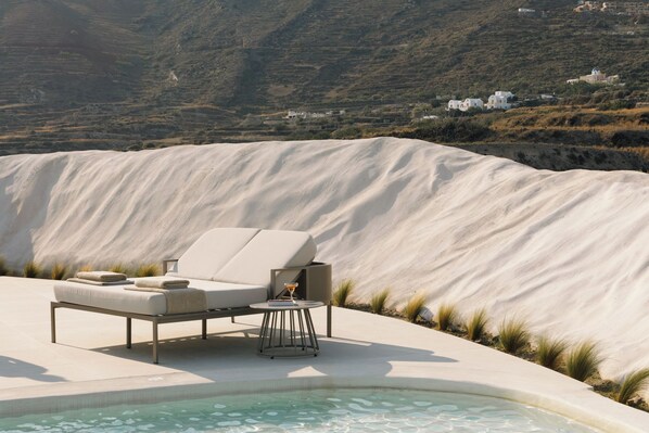 A pool area with a lounge chair and a table, surrounded by a sandy landscape and distant buildings. - Elysian Retreat (Santorini)