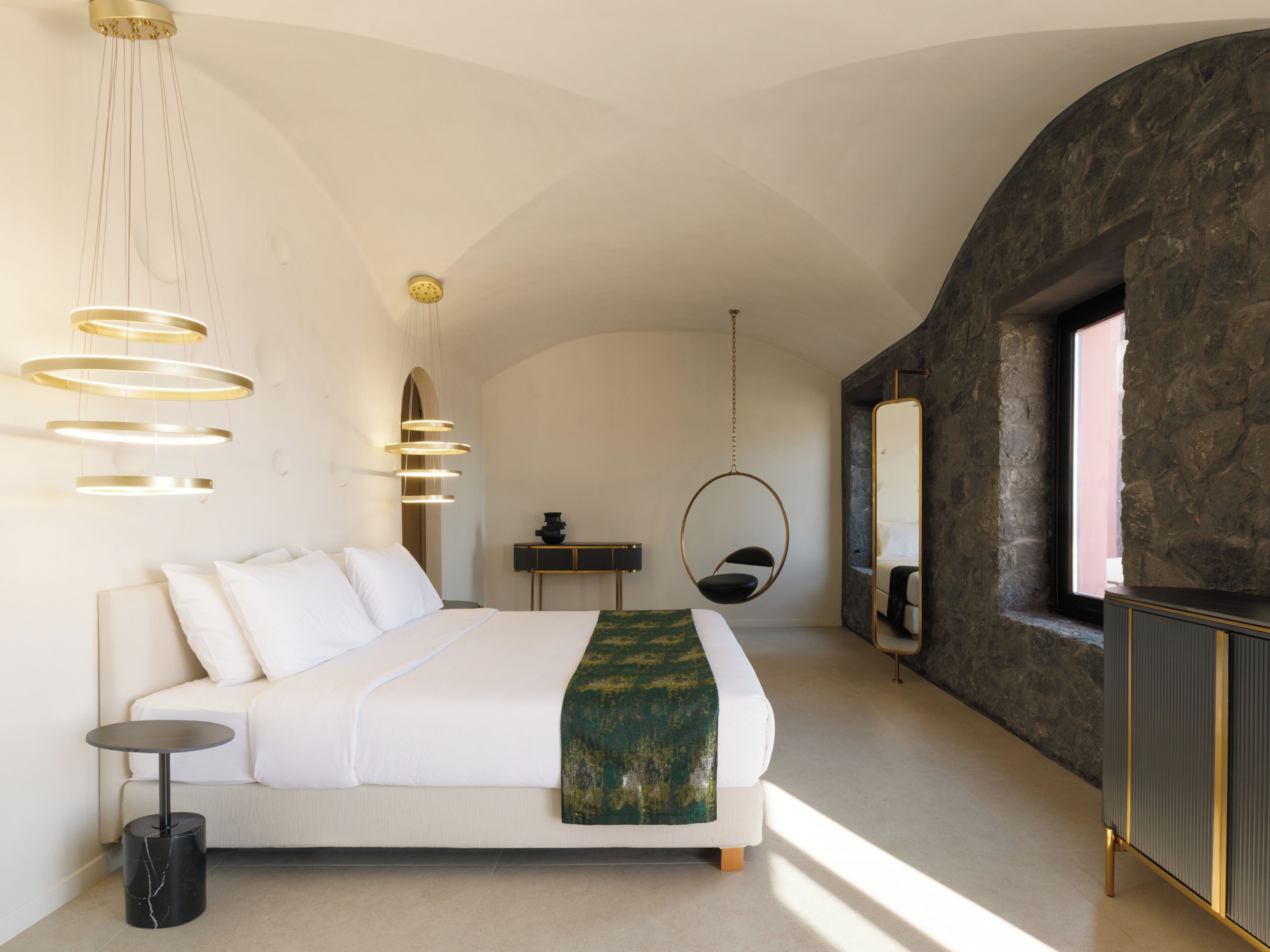 A modern bedroom with a large bed, a hanging chair, a round mirror, and a stone wall.