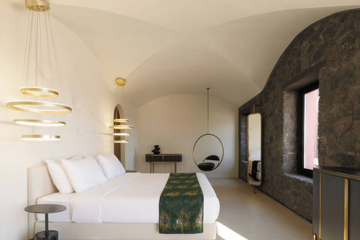 A modern bedroom with a large bed, a hanging chair, a round mirror, and a stone wall.