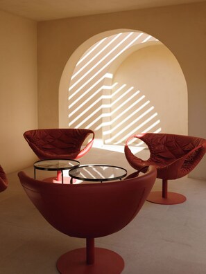 A modern interior with red chairs and a glass table, a large arched window casting light patterns, and a plain wall. - Elysian Retreat (Santorini)