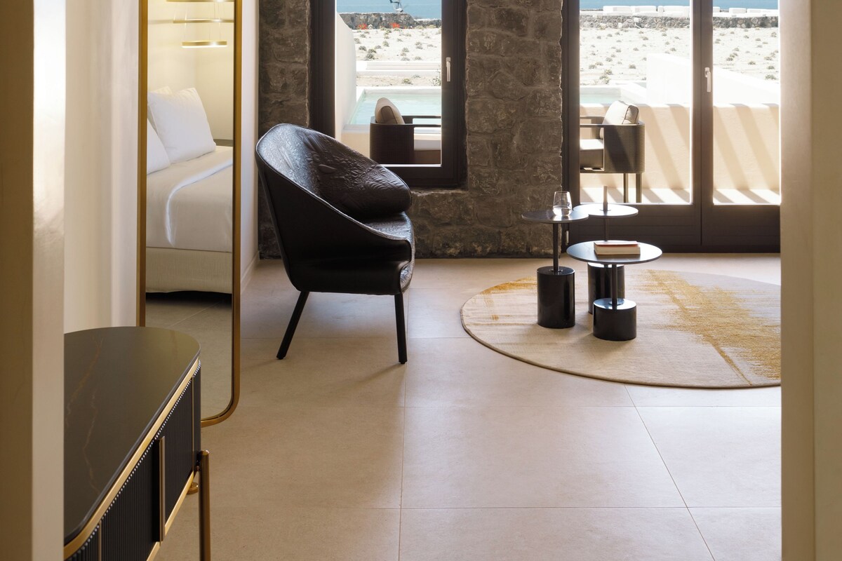 Ein Schlafzimmer mit Bett, Stuhl, Tisch und Blick auf den Strand durch ein Fenster.
