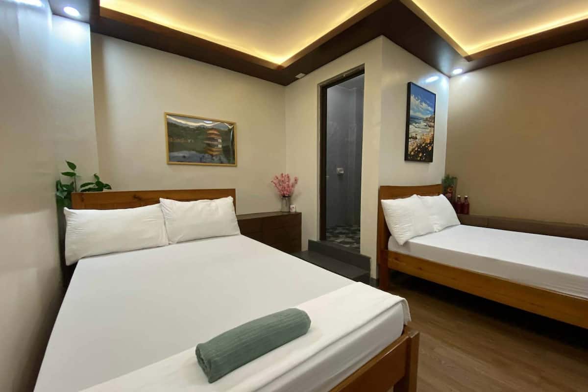 A hotel room with two beds, a door, a small table with a vase, and a painting on the wall.