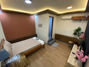 A hotel room with a bed, a desk, a chair, a small table, a potted plant, and an air conditioning unit.