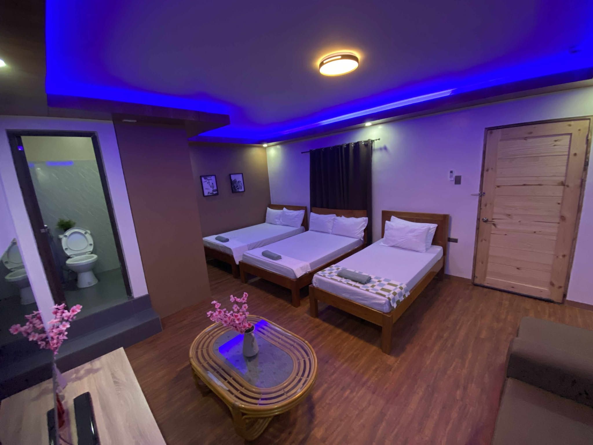 A hotel room with two single beds, a wooden floor, a small round table with a vase of flowers, and a bathroom visible through a glass partition.