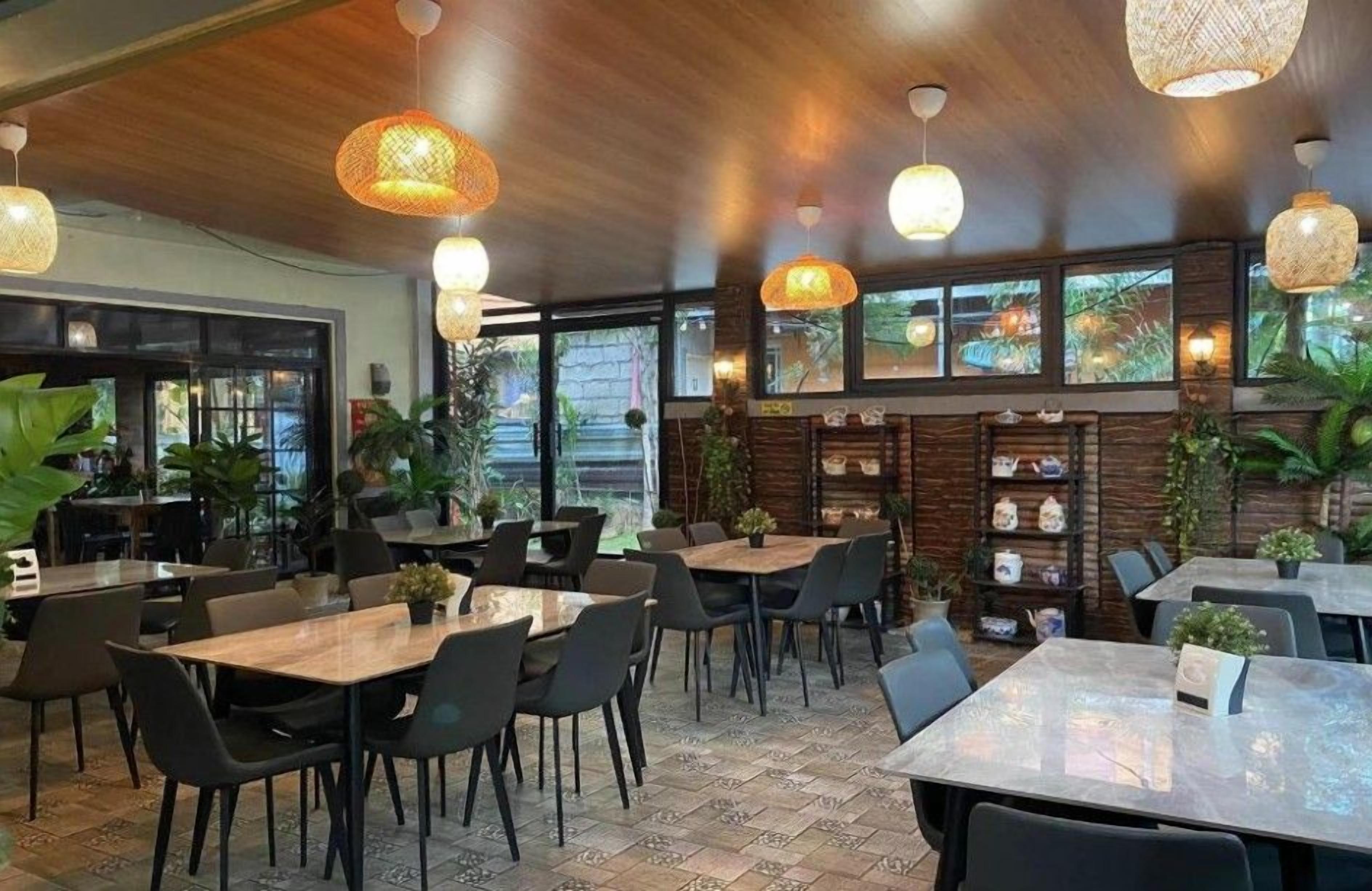 Een restaurant met houten tafels en stoelen, hangende rieten lampen, en grote ramen met uitzicht op groen.