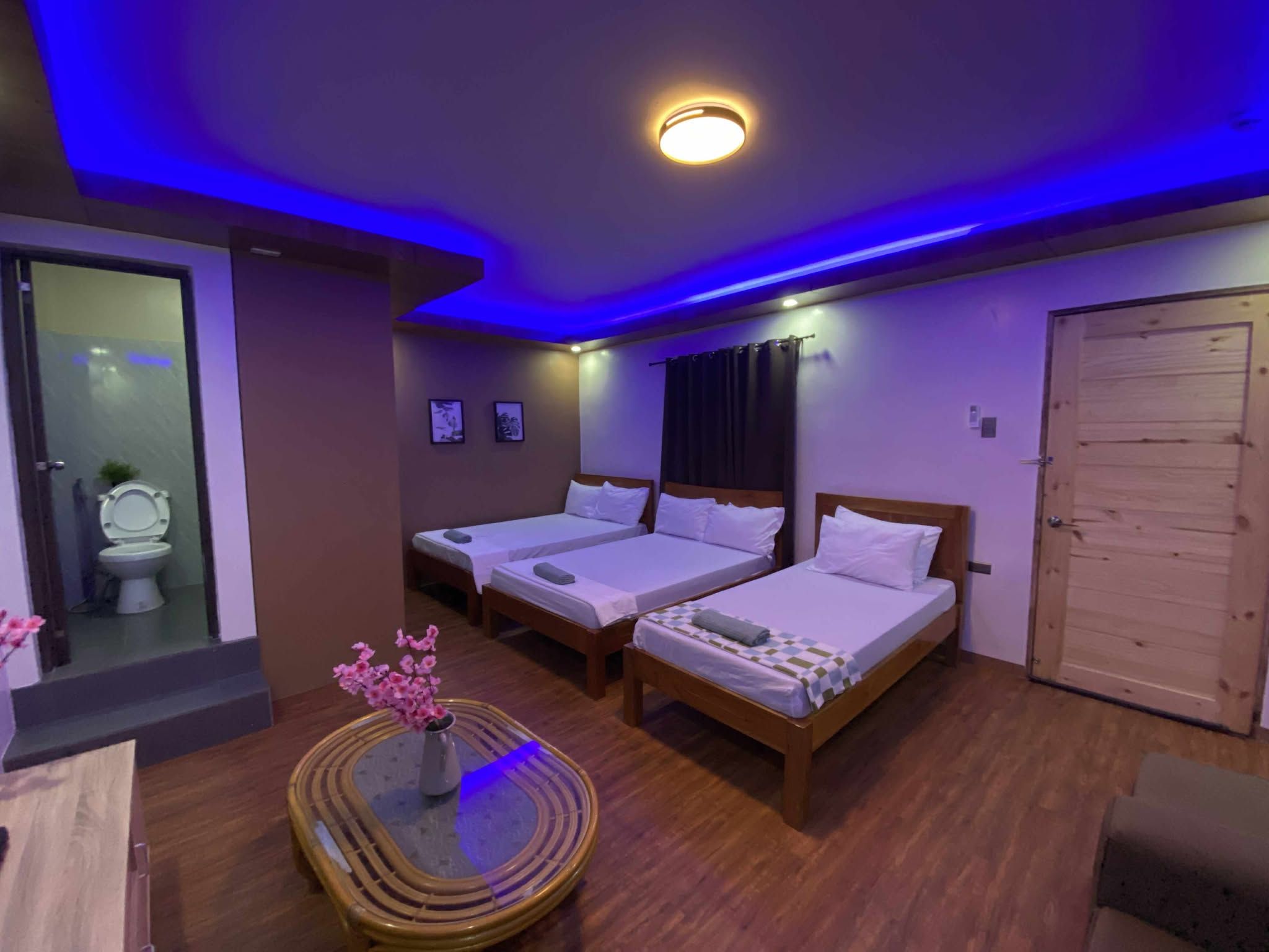 A hotel room with two single beds, a wooden door, a small table with a vase, and a bathroom visible through a glass partition.