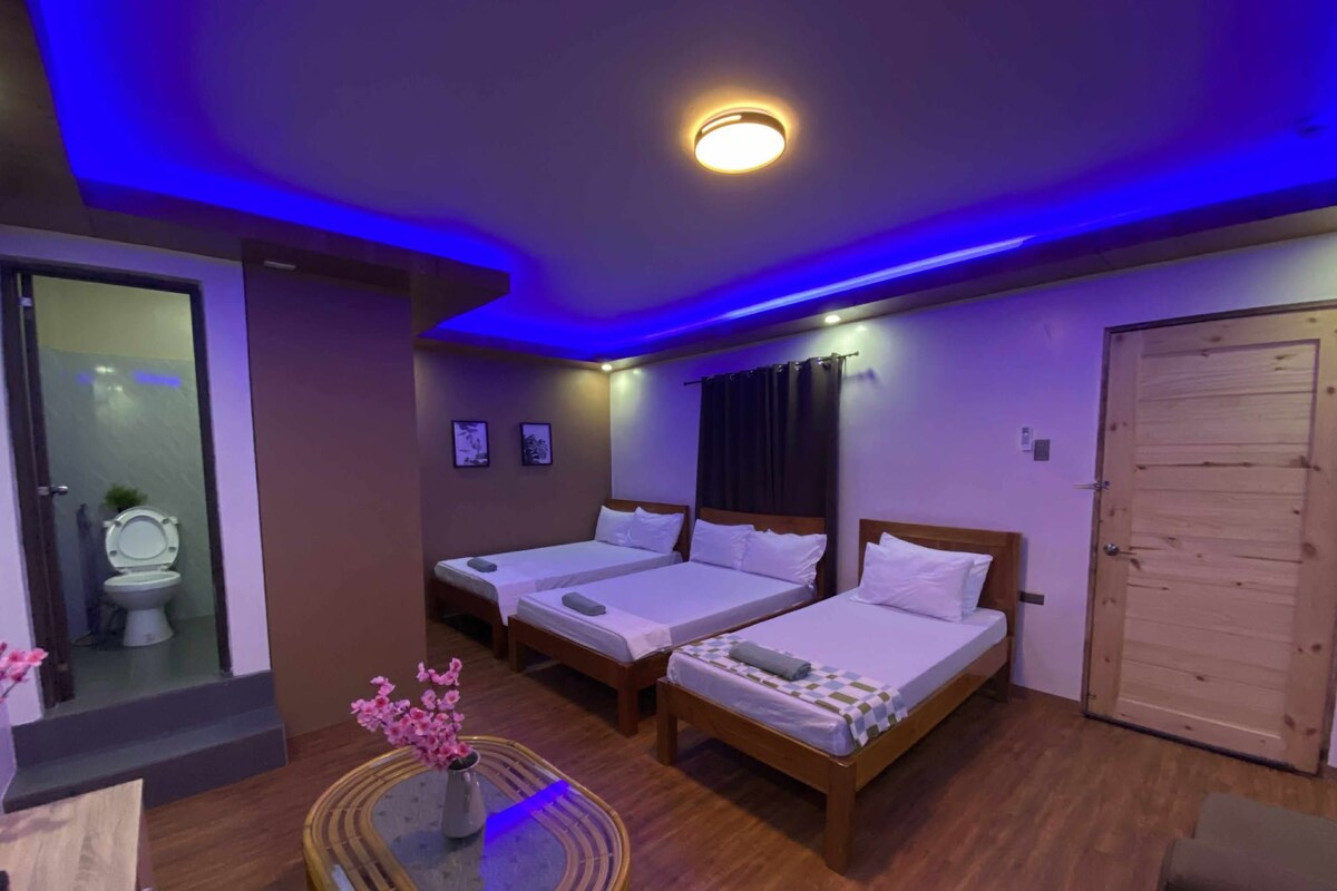 A hotel room with two single beds, a wooden door, a small table with a vase, and a bathroom visible through a glass partition.