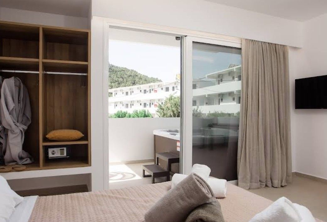 A hotel room with a bed, a wardrobe, a sliding glass door to a balcony, and a TV mounted on the wall.