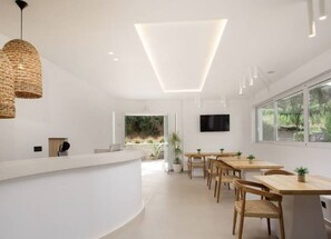 Un intérieur moderne avec un comptoir d’accueil incurvé, des tables en bois, des suspensions en osier, un téléviseur à écran plat et de grandes fenêtres.