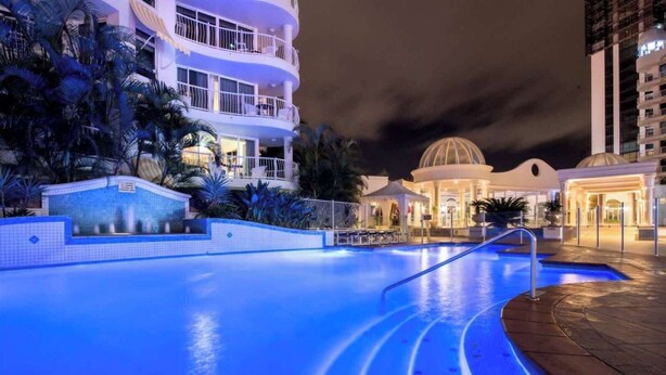 A nighttime swimming pool with a curved edge, surrounded by a landscaped area with palm trees and a building with balconies. - The Phoenician Luxe Suite (Broadbeach)