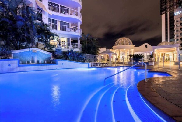 Ein Pool bei Nacht mit gewölbter Kante, umgeben von einer angelegten Grünfläche mit Palmen und einem Gebäude mit Balkonen.
