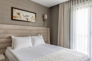 Habitación de hotel con una cama grande, cabecero, un cuadro, lámpara y ventana con cortinas.