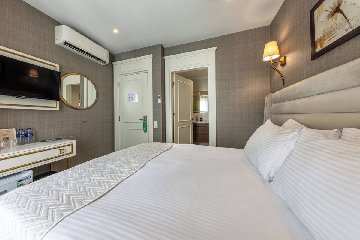 A hotel room with a bed, a desk with a mirror, a television, and an air conditioning unit.
