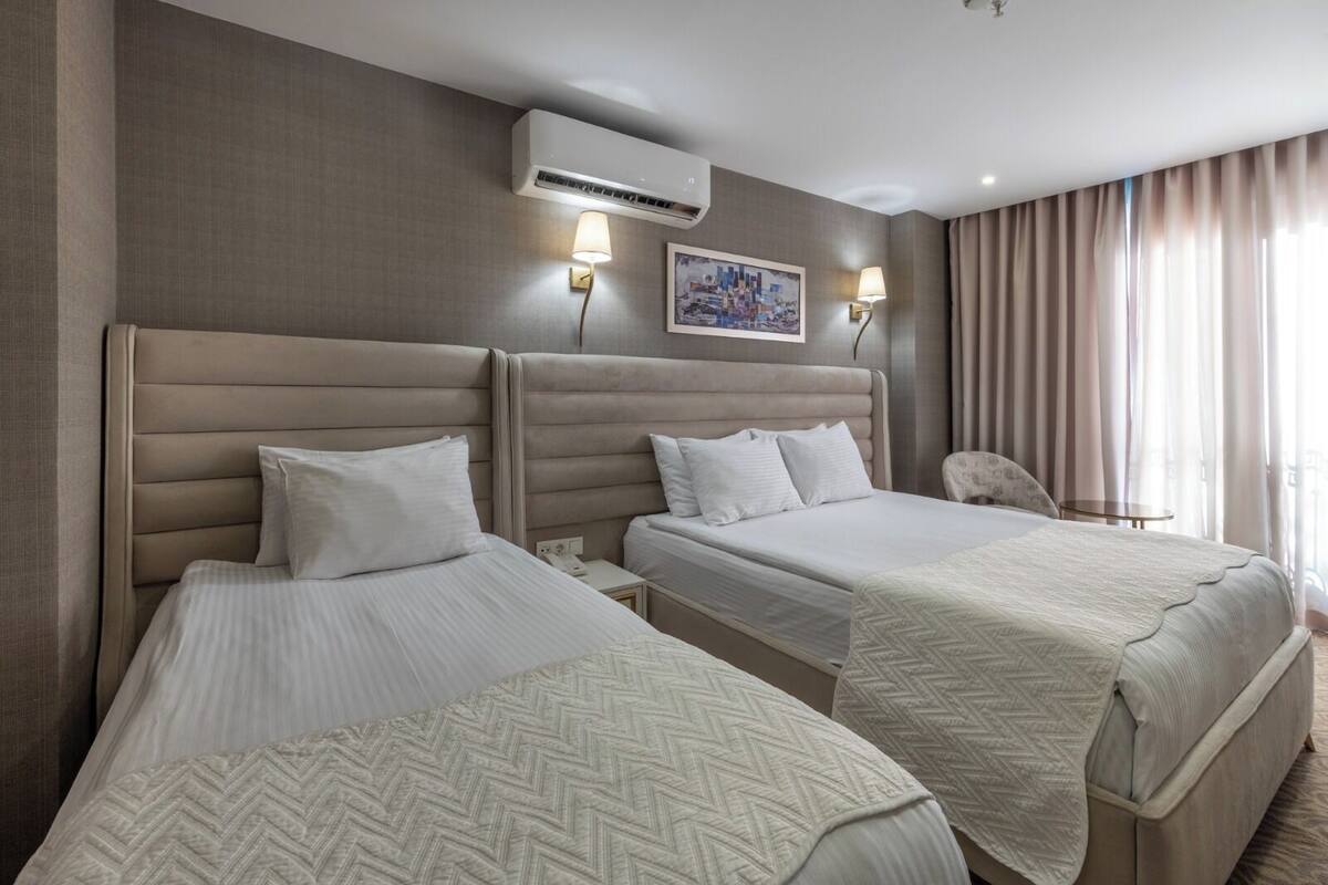 Ein Hotelzimmer mit zwei Betten, einer Wand-Klimaanlage, zwei Wandleuchten, einem Gemälde und einem Sessel.