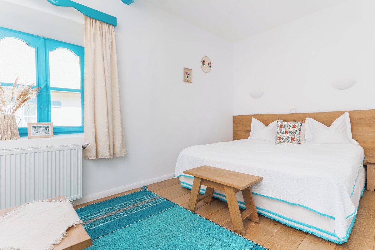 A bedroom with a bed, a wooden nightstand, a window with blue shutters, a radiator, and a wall-mounted picture.
