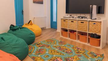 A room with a large flat-screen TV mounted on a white cabinet, bean bags in green and orange, a colorful patterned rug, and a blue door.