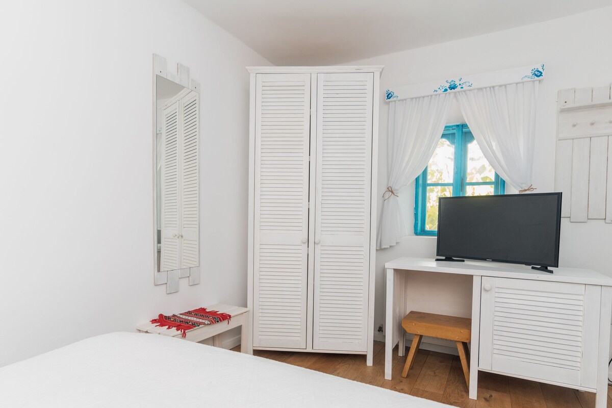 A bedroom with a bed, a desk with a TV, a wardrobe, and a window with curtains.