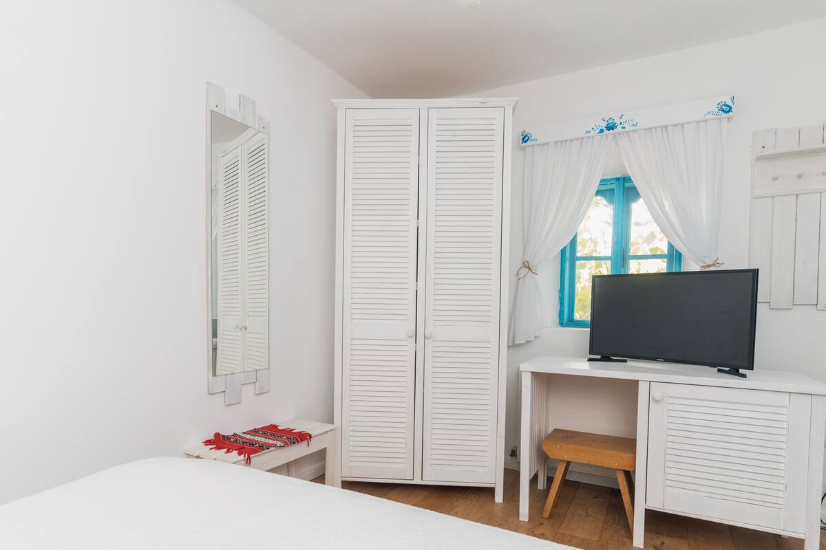 A bedroom with a bed, a desk with a TV, a wardrobe, and a window with curtains.