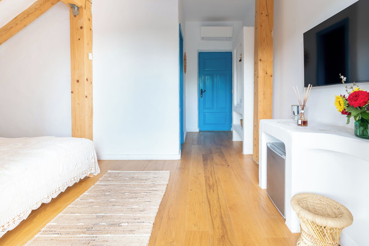 A bedroom with a wooden ceiling, a white bed, a woven rug, a blue door, and a small wicker stool.