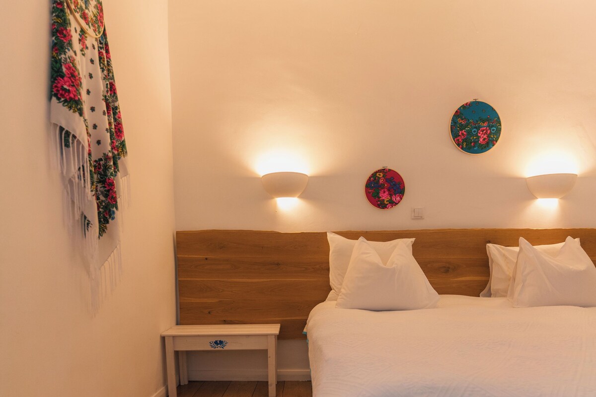 A bedroom with a wooden headboard, a white bedspread, two wall-mounted lights, a small bedside table, and a decorative wall hanging.