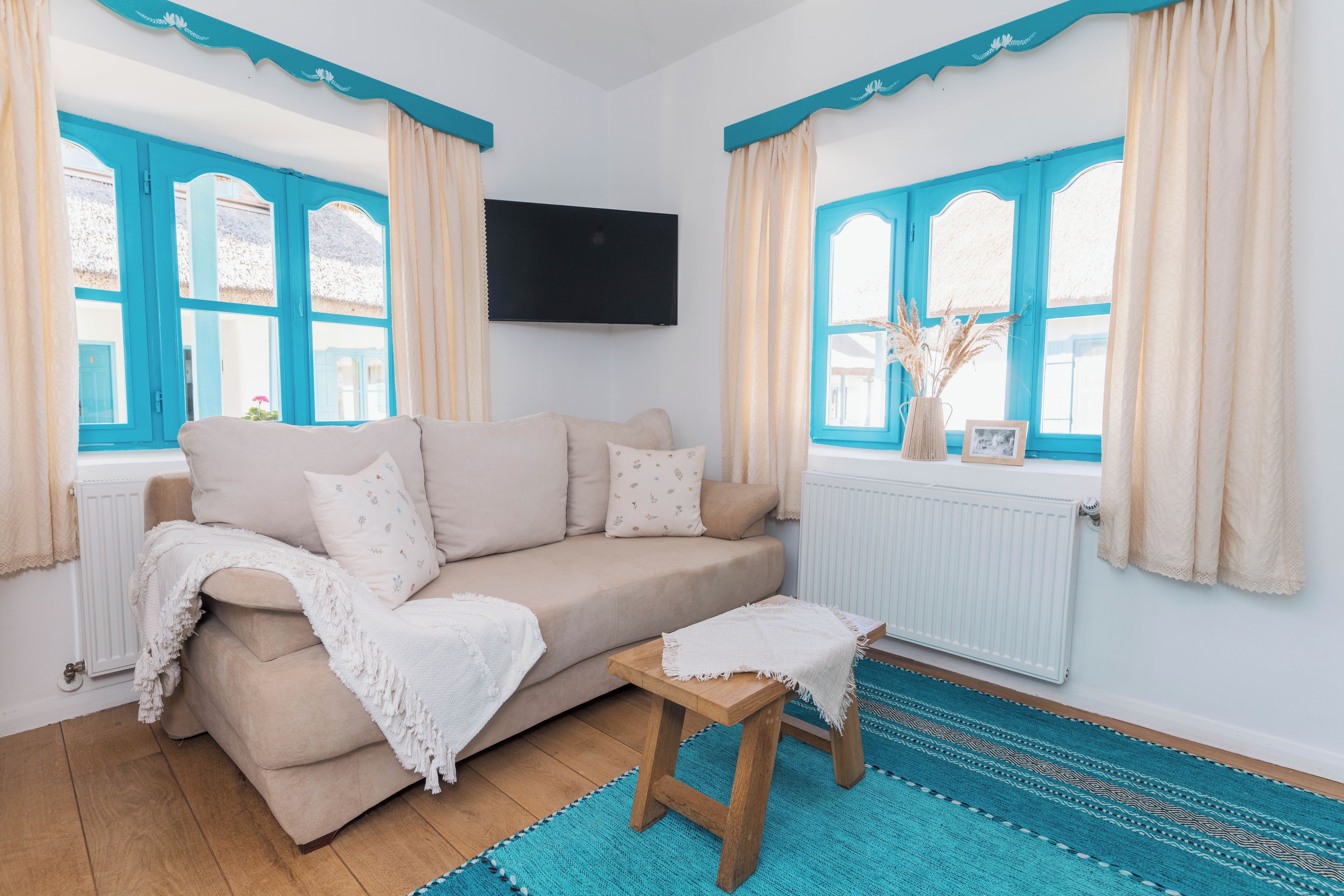 A living room with a beige sofa, a small wooden stool, a blue patterned rug, and a TV mounted on the wall.