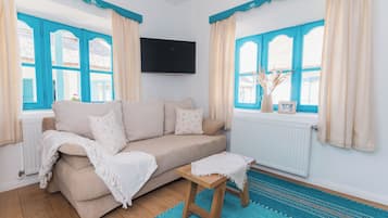 A living room with a beige sofa, a small wooden stool, a blue patterned rug, and a TV mounted on the wall.