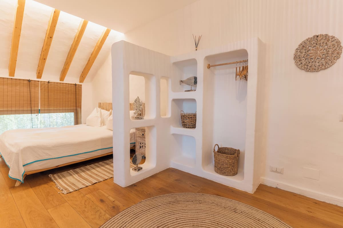 A bedroom with a bed, wooden beams, a built-in shelf with baskets, and a decorative wall piece.